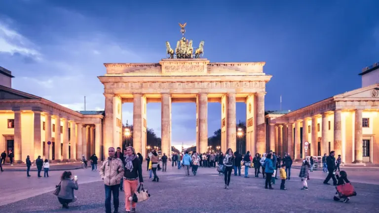 Das beleuchtete Brandenburger Tor in Berlin bei Dämmerung mit vielen Passanten auf dem Platz.