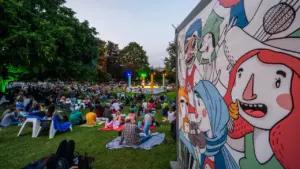 Zuschauer sitzen auf Picknickdecken im Park vor einer bunt beleuchteten Bühne bei einer Abendveranstaltung.