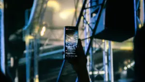 Eine Hand hält ein Smartphone hoch, das eine Bühnenveranstaltung bei Nacht filmt.