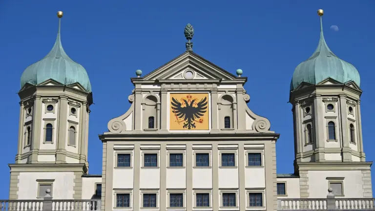 Prächtige Architekturfassade mit zwei Türmen und einem Adlerwappen vor einem strahlend blauen Himmel.