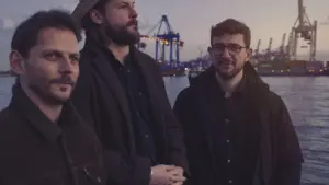 Drei Männer stehen vor einer Hamburger Hafen-Kulisse mit Kränen bei einsetzender Abenddämmerung am Wasser.