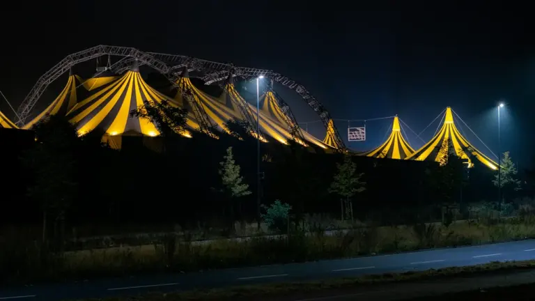 Ein großes, beleuchtetes Zirkuszelt bei Nacht in einer städtischen Umgebung.