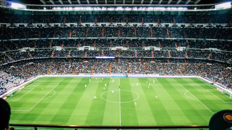 Weitwinkelaufnahme eines vollen Fußballstadions unter Flutlicht während eines spannenden Spiels am Abend.