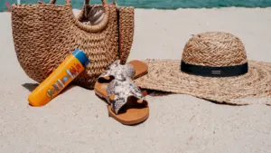 Strandzubehör mit Sonnencreme, Strohhut und Tasche im Sand bei sommerlicher Hitze.