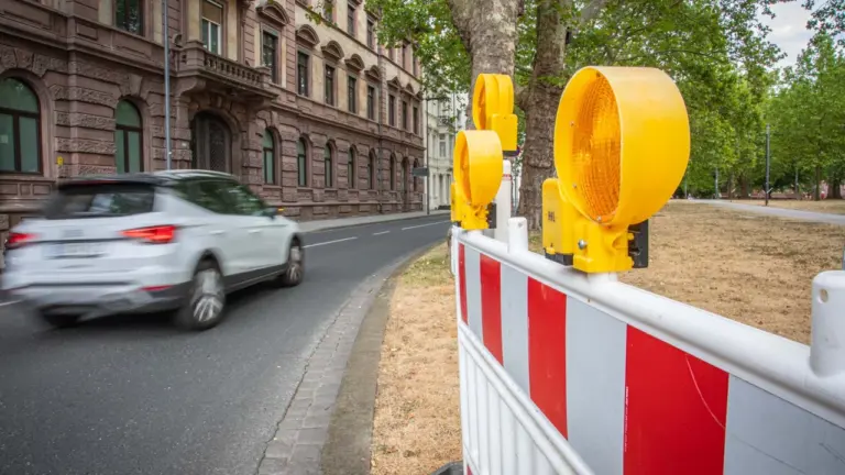 Ein weißes Auto fährt schnell an einer rot-weißen Baustellenabsperrung mit gelben Warnleuchten vorbei.