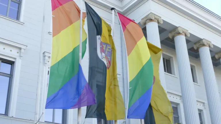 Regenbogenfahnen und eine Flagge mit Wappen wehen vor einem klassischen Gebäude mit Säulen.