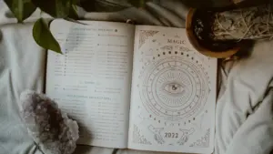 Ein offenes Tagebuch mit astrologischen Zeichnungen, einem Kristall und Pflanzen auf einer weichen Decke.