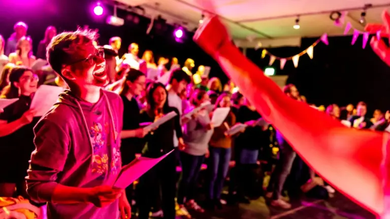 Ein lachender junger Mann singt in einem Chor auf einer Bühne mit violetter Beleuchtung.