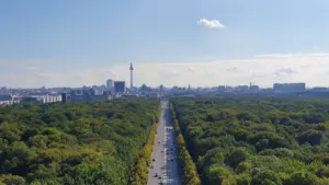 Blick von der Berliner Siegessäule auf eine von Bäumen gesäumte Straße und Berliner Skyline.